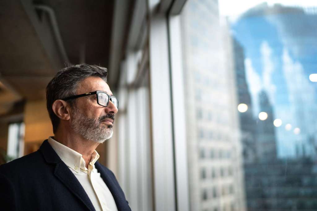 Man wearing glasses and a suit looks out of a large window in an office building.