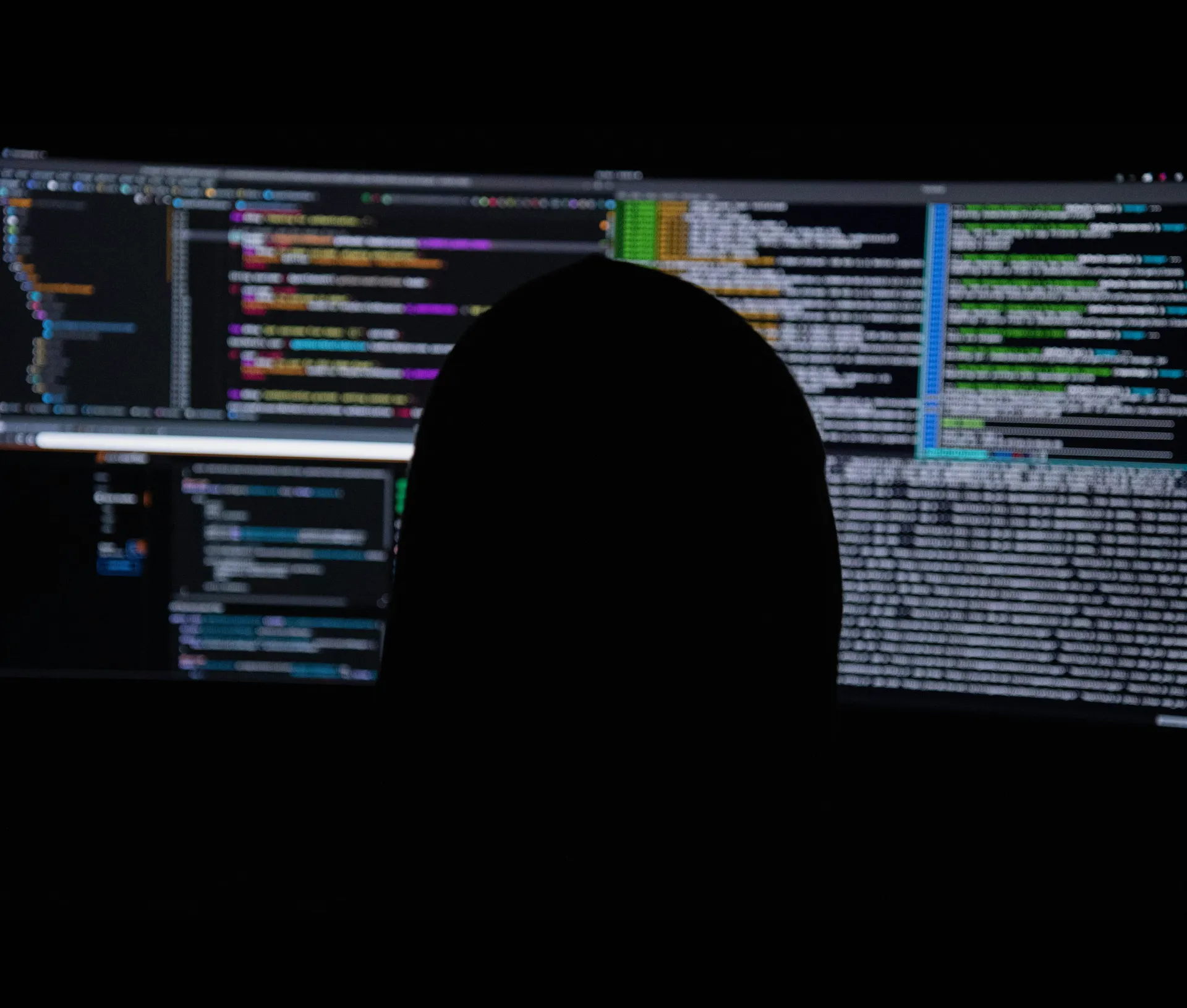 A silhouette of a person sits in front of multiple computer monitors displaying colourful lines of code in a dark room, suggesting programming or hacking activity—perhaps highlighting the growing role of AI and its impact on UK businesses confronting cyber threats from sophisticated criminals.