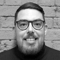 A man with glasses, short dark hair, and a beard smiles in front of a brick wall.