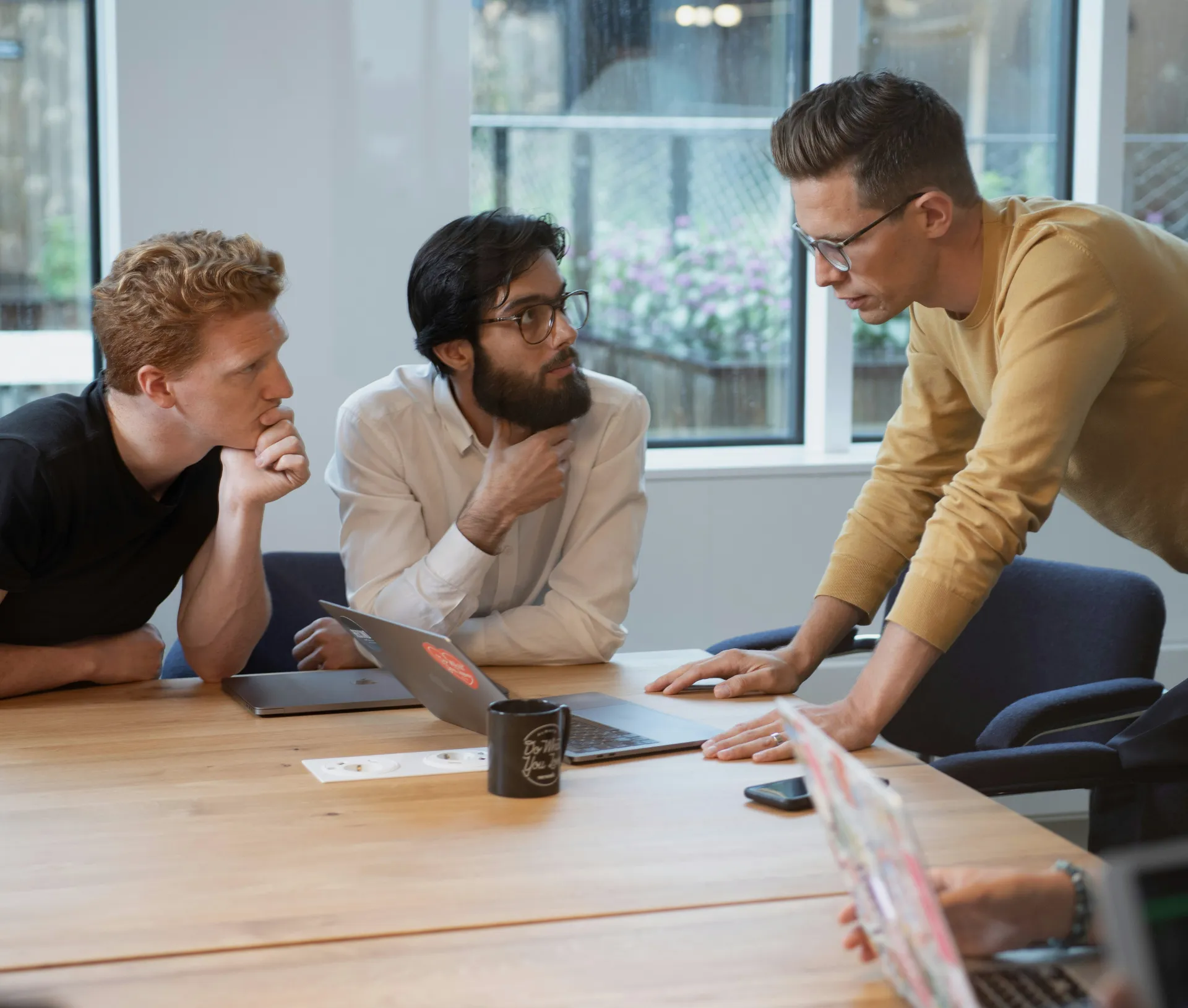 Three people sit at a conference table, two listening intently while one stands and gestures towards a laptop during an IT policy review. A coffee mug and papers are on the table; large windows show a blurred outdoor view, setting the scene for more than just a set and forget approach to the annual IT policy.