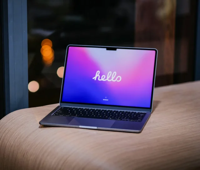 A laptop with a colourful screen displaying the word "hello" in cursive sits on a light wooden surface near a window, with blurred lights visible in the background—an inviting scene familiar to many UK users setting up their personal Apple Accounts or exploring AppleCare One cover.