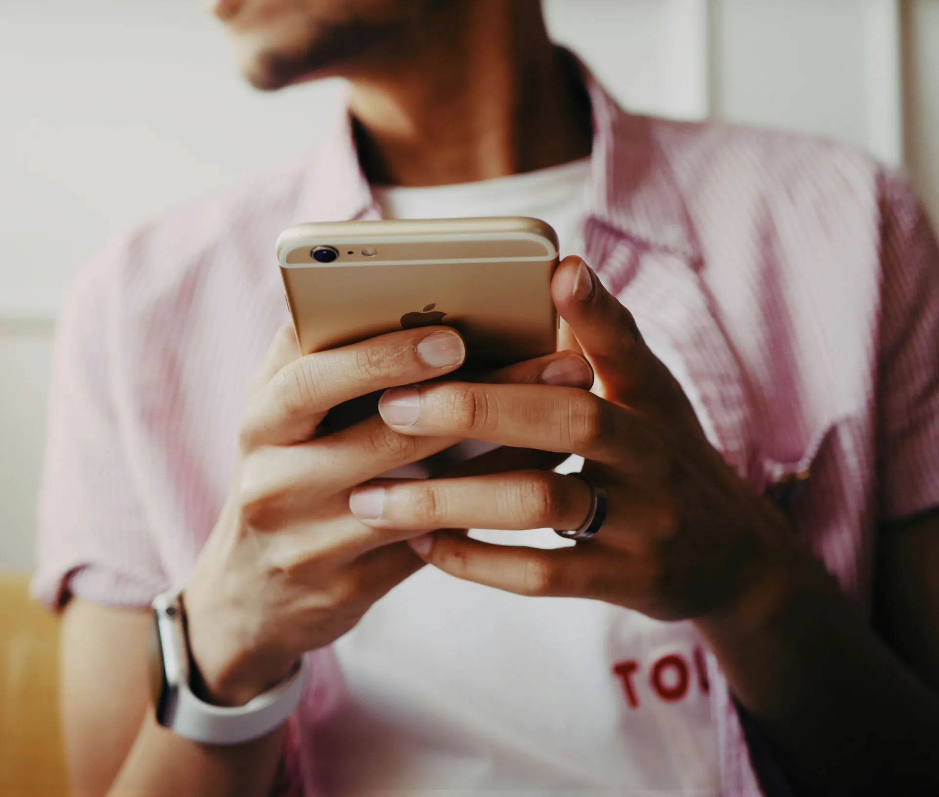 A person wearing a pink shirt holds a gold Apple iPhone with both hands. They are wearing a watch and a ring, and their face is partially out of focus, highlighting the seamless blend of work and personal data separation that BYOD iPhones offer.