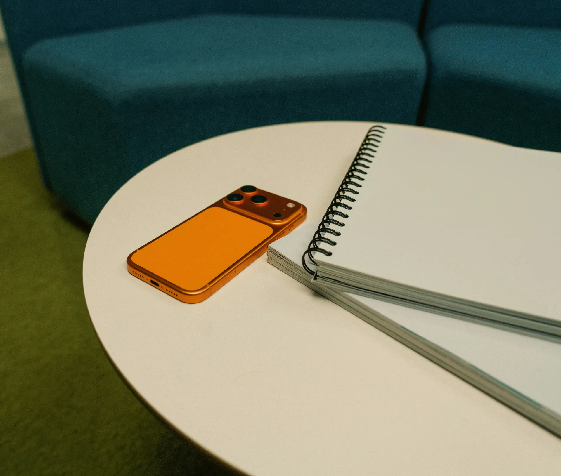 An orange mobile phone and an open spiral notebook rest on a round white table, with a blue sofa and green carpet in the background—an ideal setting for those using BYOD iPhones to maintain work–personal data separation.