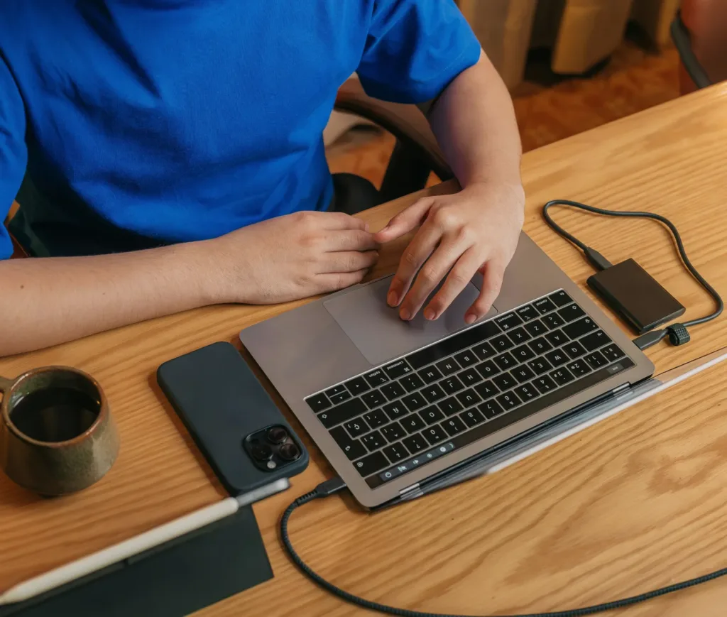 A person in a blue shirt sits at a wooden desk with a laptop, mobile phone, external drive, pen, notepad, and a mug. The person’s hands rest on the laptop’s keyboard as they focus on backup restore testing to ensure seamless business data recovery.