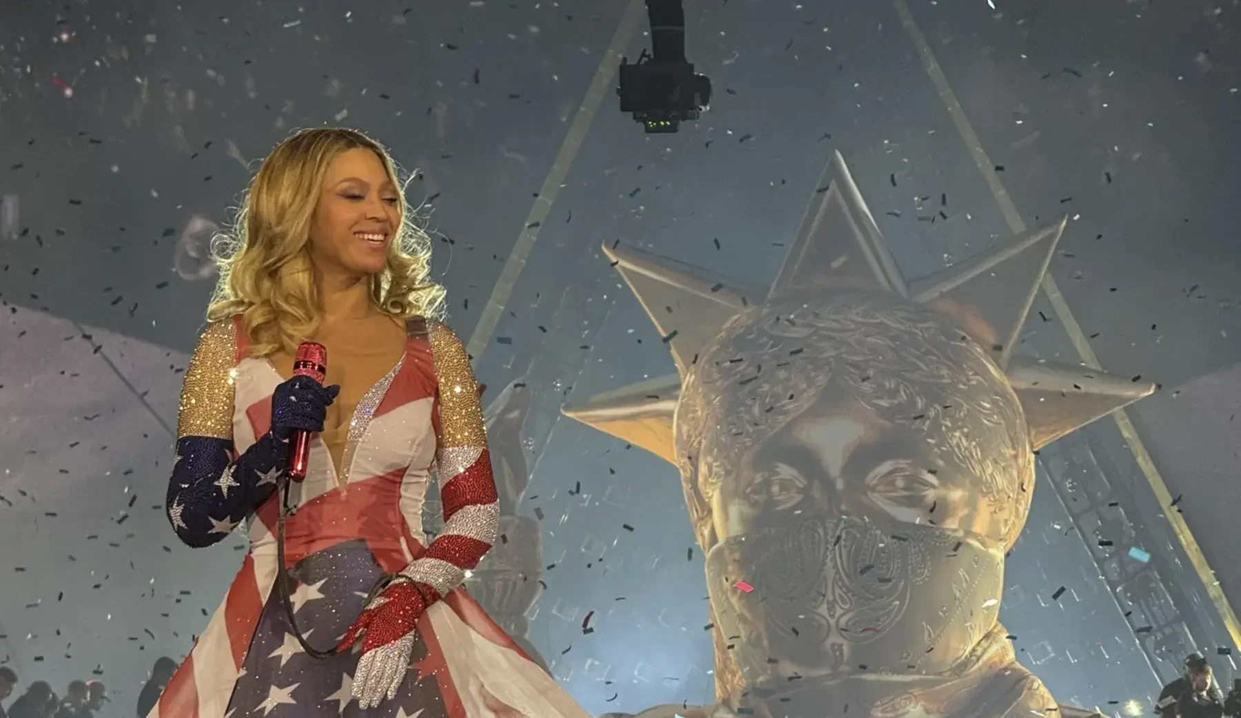 A woman in a star spangled outfit, channeling beyoncé, holds a microphone on stage beside a large metallic sculpture as confetti falls around them.