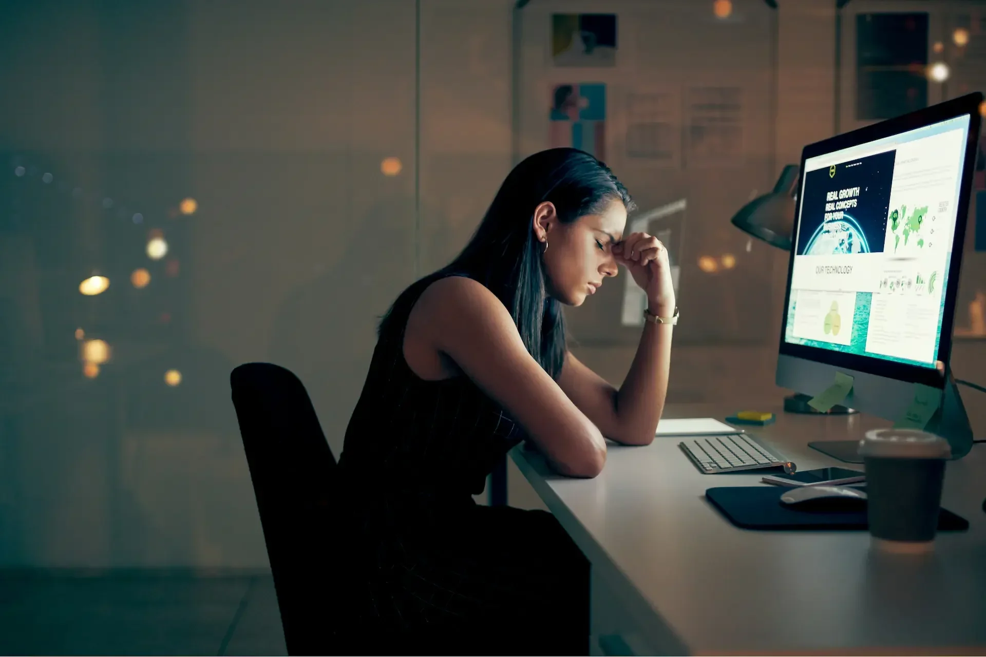 A woman sits at a desk in a dimly lit office at night, looking stressed as she stares at charts and data on her computer—signs of burnout from long hours managing a demanding it setup.
