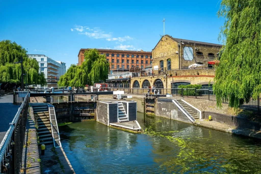 Canal with a lock, surrounded by brick buildings and greenery on a sunny day.