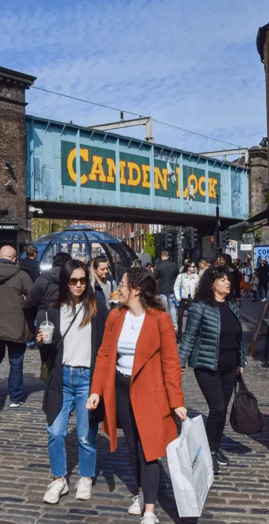 People walking under the iconic Camden Lock railway bridge in London on a clear day, just a stone's throw from the bustling vibes of Camden Town and its famous brewery.