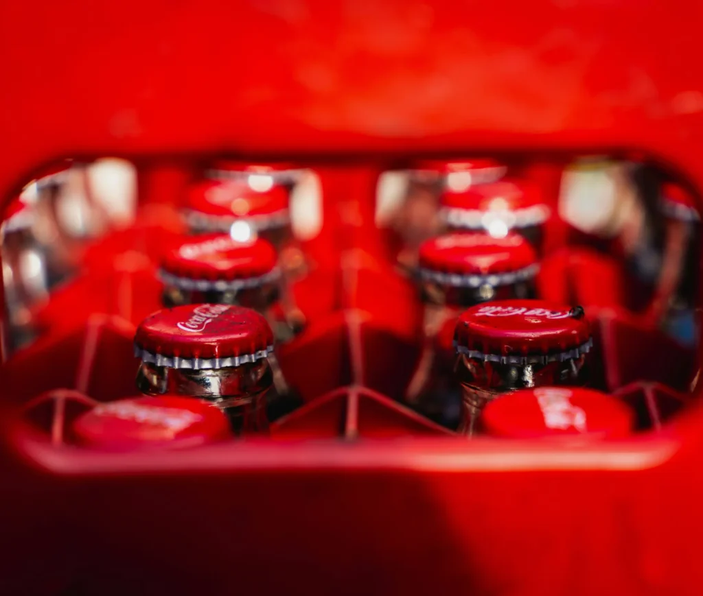 Close up of glass Coca Cola bottles with red caps in a red crate, viewed from above. The image is vibrant with a dominant red colour, highlighting the iconic branding and bottle tops—an immediately recognisable scene akin to the signature packaging seen across leading UK milk production brands like Fairlife.