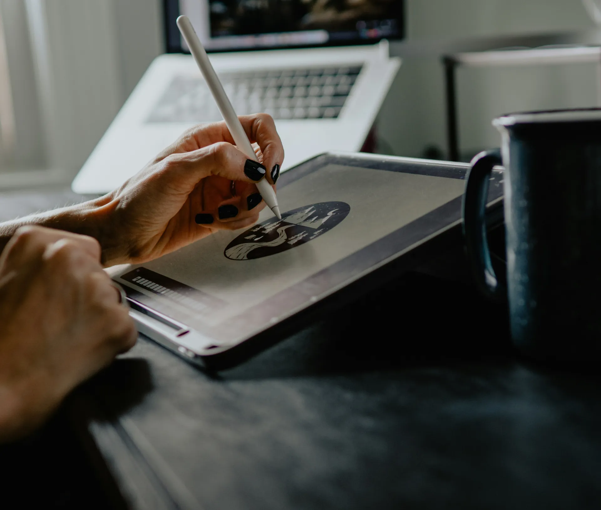 A creative draws on a digital tablet with a stylus, creating a black and white image. A laptop and a coffee mug are visible in the background on a dark desk, suggesting an inspiring workspace for creatives perfecting their profile.
