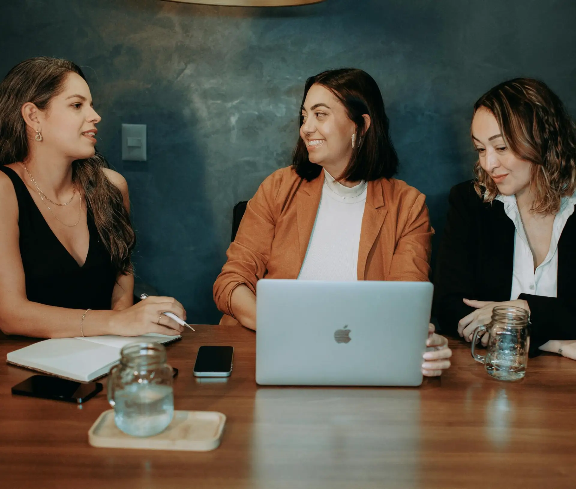 Three women sit at a table with a laptop, notebook, phone, and water glasses, engaged in conversation about reducing burnout and deep work in a modern office setting with a dark blue background.