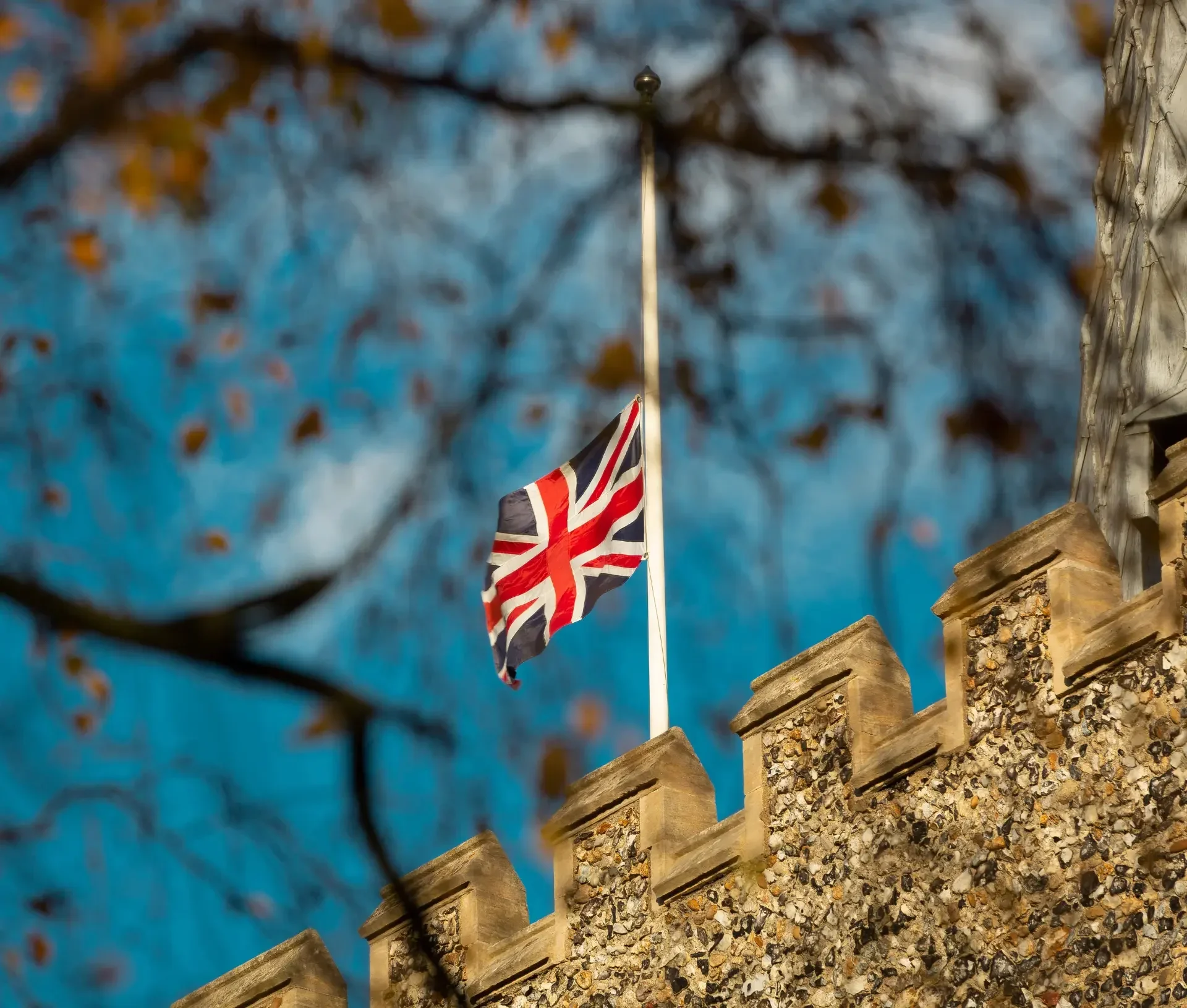 Union jack flag at half mast on a stone building, with tree branches and autumn leaves in the foreground against a blue sky—a reminder for uk businesses to focus on cyber resilience as 2025 approaches.