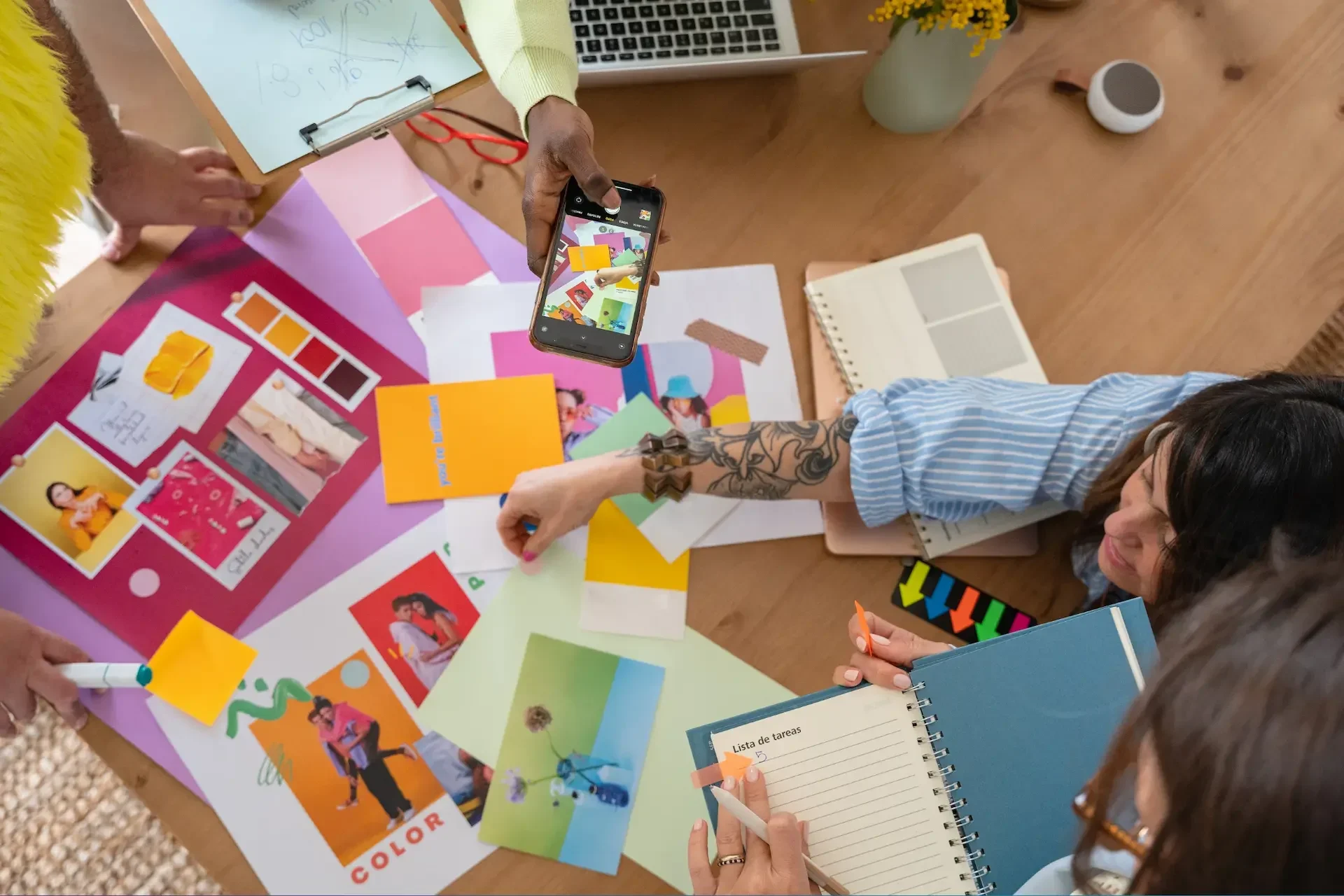 Four people collaborate at a table covered with colorful papers, photos, notebooks, and color swatches; one person takes a photo with a smartphone, ready to share it instantly using collaboration and file sharing services.