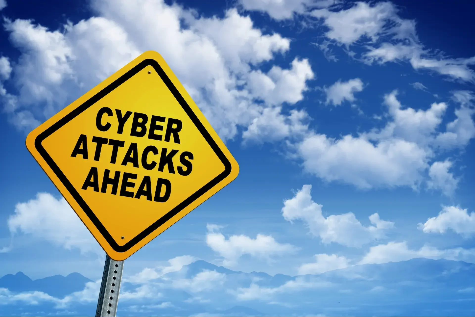 Yellow road sign with the words "cyber attacks ahead" against a blue sky with clouds, highlighting the risks uk businesses and smes face from cyber attacks.