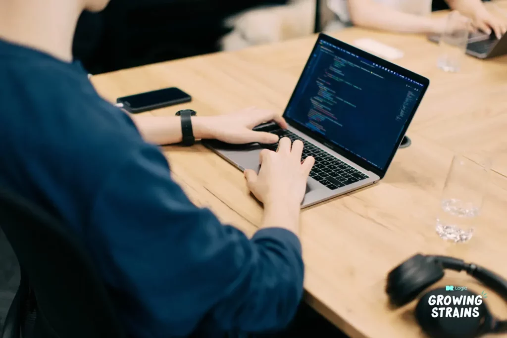 Person typing code on a laptop at a wooden desk with a smartphone nearby.