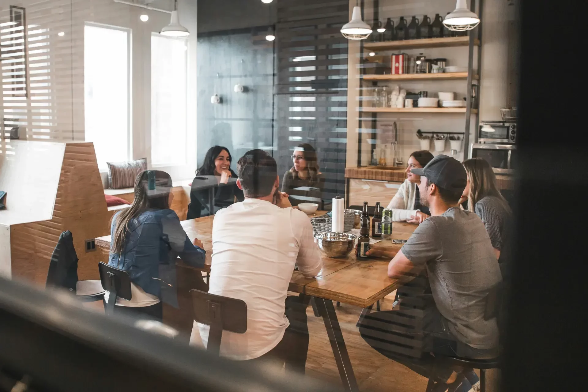 A group of people are sitting around a wooden table in a modern office setting, engaging in a discussion.
