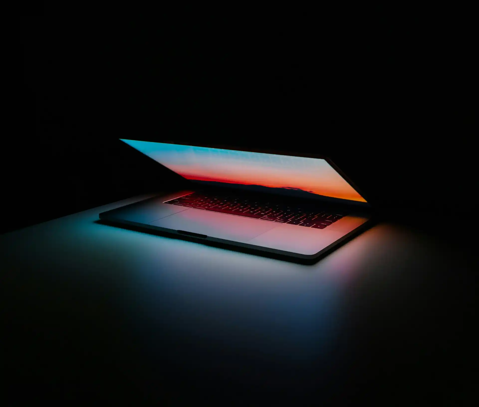 A partially closed laptop glows in a dark room, with light from its colourful screen reflecting softly on the surface below—a quiet reminder of the importance of dark web monitoring to prevent breach and exposure.