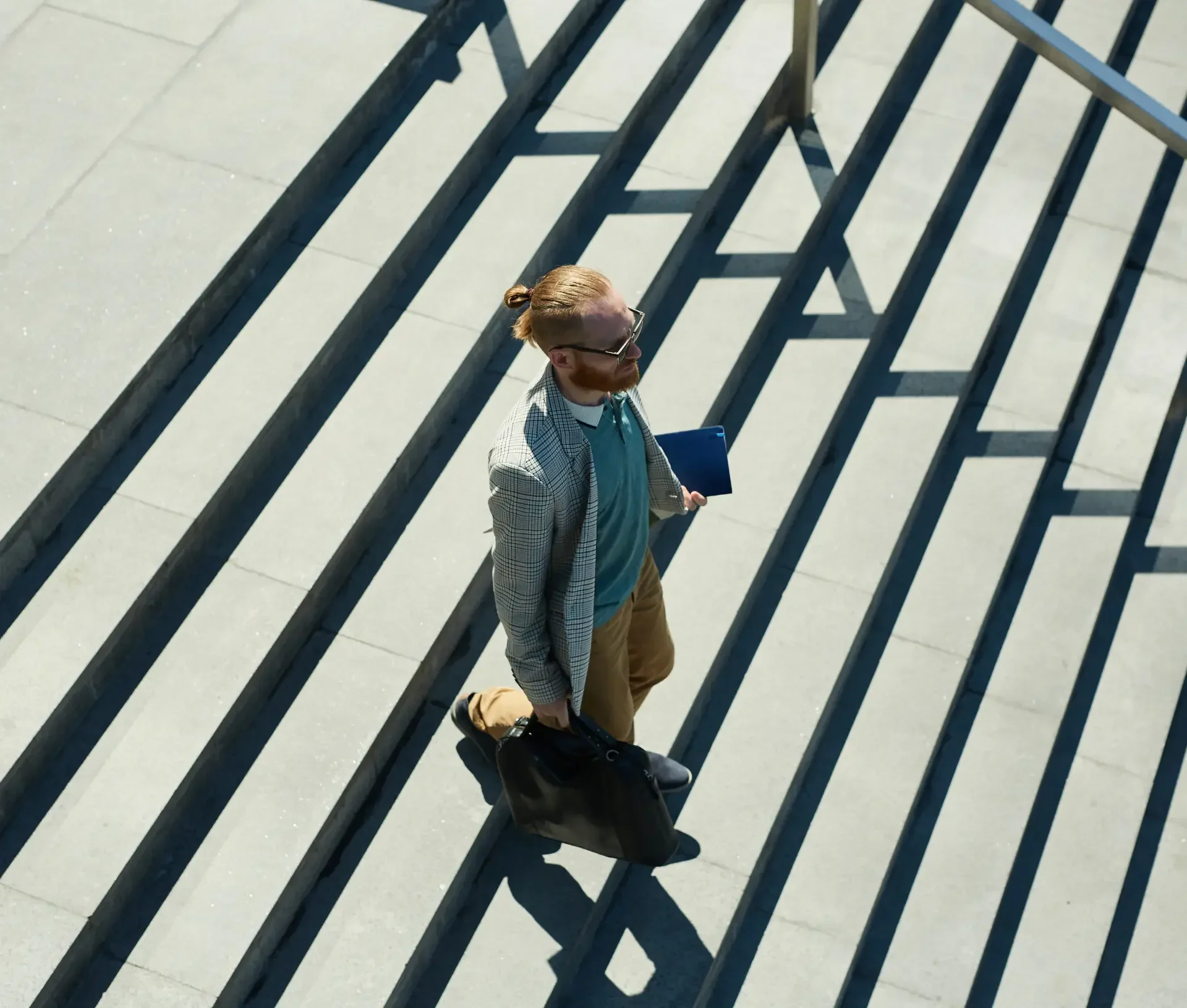 Man with a beard and glasses walks up outdoor concrete steps holding a black bag and a blue folder, dressed in a blazer and light trousers—perhaps contemplating offboarding security or potential security gaps within the company.