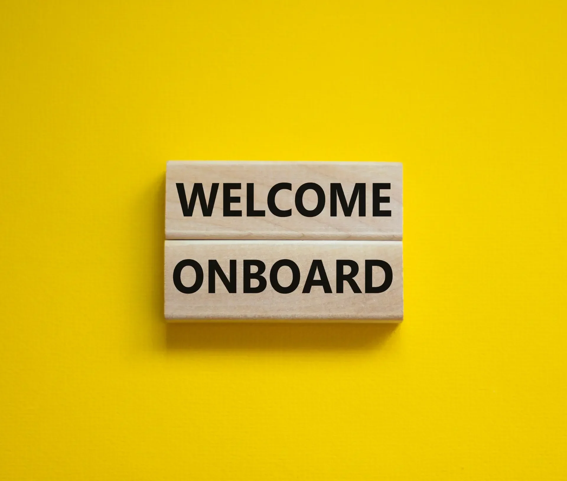 Two wooden blocks with "welcome" and "onboard" printed in black sit on a bright yellow background, symbolizing a warm start to employee onboarding for any it manager’s checklist.
