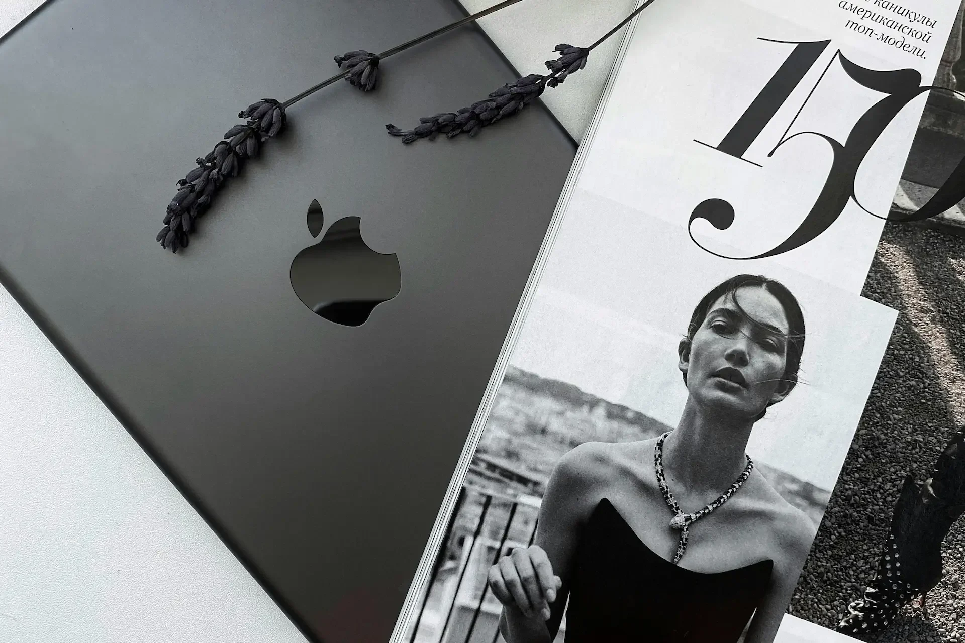 A closed macbook with a lavender sprig next to a monochrome fashion magazine showing a woman in a necklace and black dress.