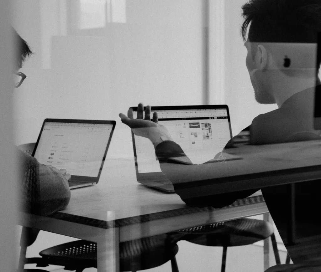 Two people sit at a table using laptops, engaged in discussion—perhaps reviewing the details of a DMS platform migration or considering whether to renegotiate their IT support contract. One faces the camera, gesturing with a hand, whilst the other looks at their screen. The scene is black and white, viewed through glass.