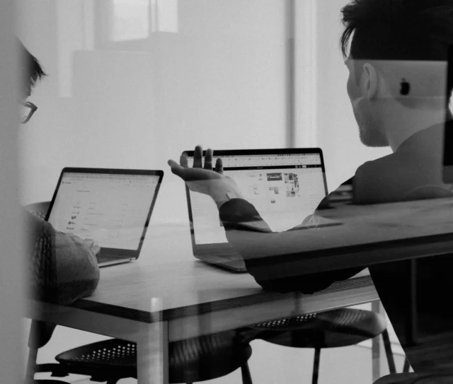 Two people sit at a table using laptops, engaged in discussion—perhaps reviewing the details of a DMS platform migration or considering whether to renegotiate their IT support contract. One faces the camera, gesturing with a hand, whilst the other looks at their screen. The scene is black and white, viewed through glass.