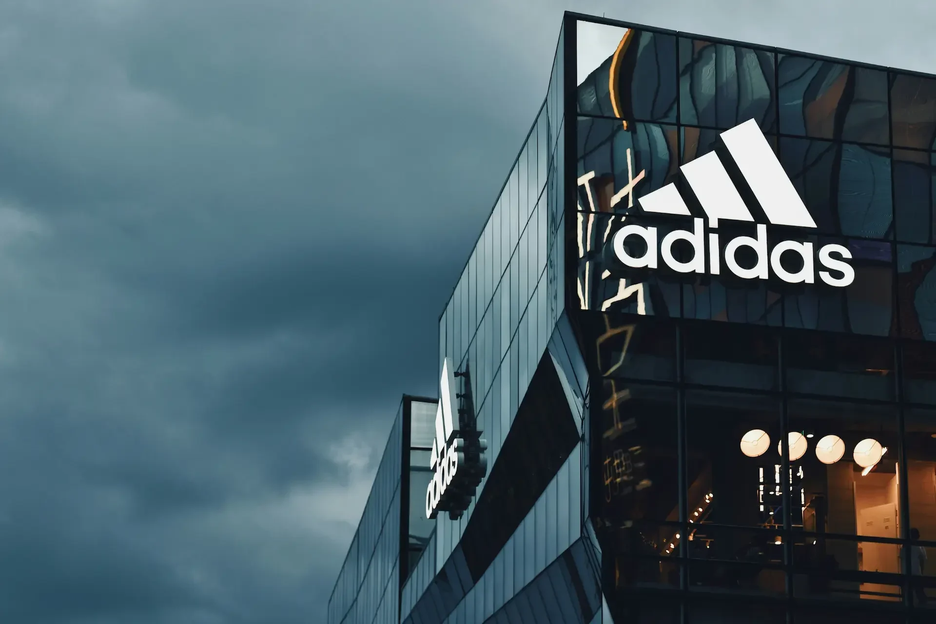 Modern glass building featuring the illuminated adidas logo and branding, set against a cloudy sky, reflects the company's commitment to brand security amid evolving cyber attack threats.
