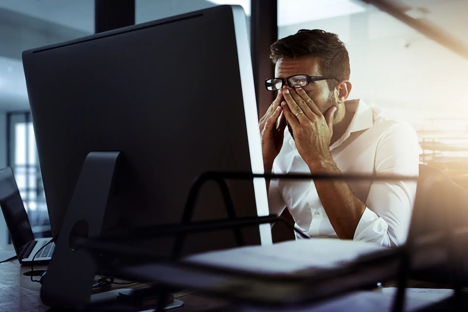 A man wearing glasses sits at a desk in front of a computer monitor, rubbing his eyes with both hands, appearing tired from working on it support tasks in an office setting.