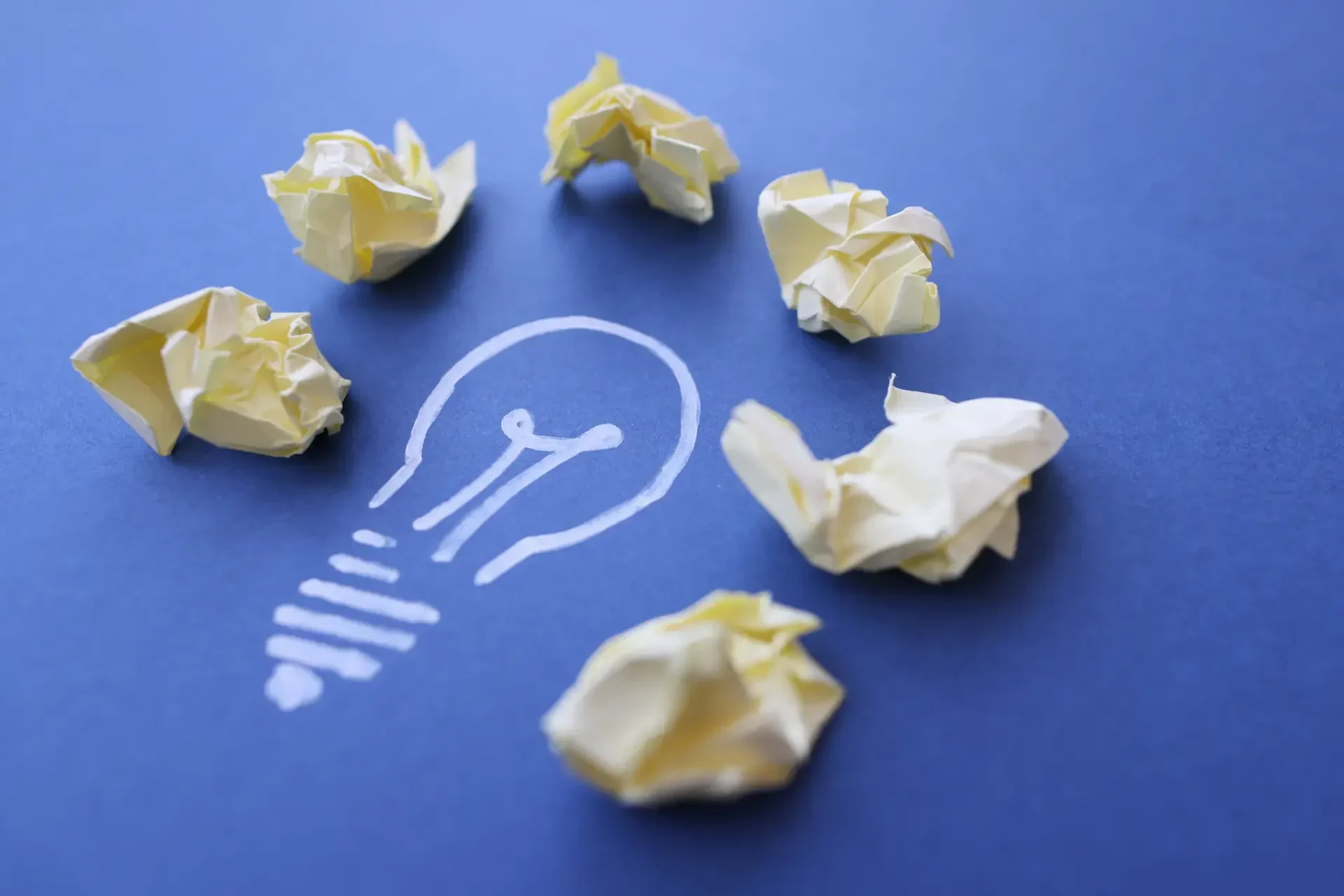 A white light bulb is drawn on a blue surface, surrounded by six crumpled yellow paper balls, symbolizing bright ideas and future proof technology solutions.