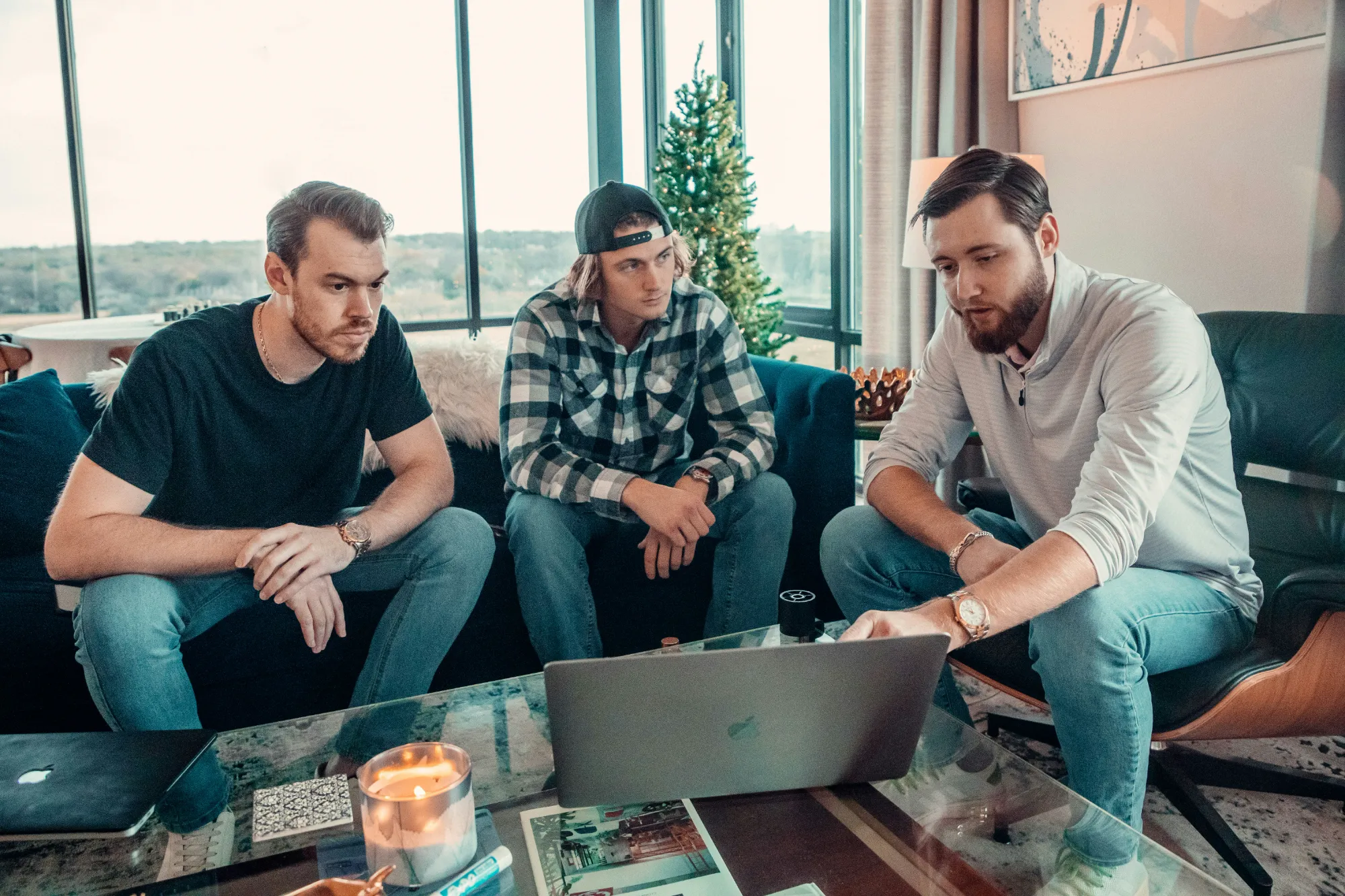 Three men sit around a coffee table in a modern living room, focused on a laptop. One gestures at the screen whilst the others look on. A candle, magazines, and a window with a scenic view are visible.