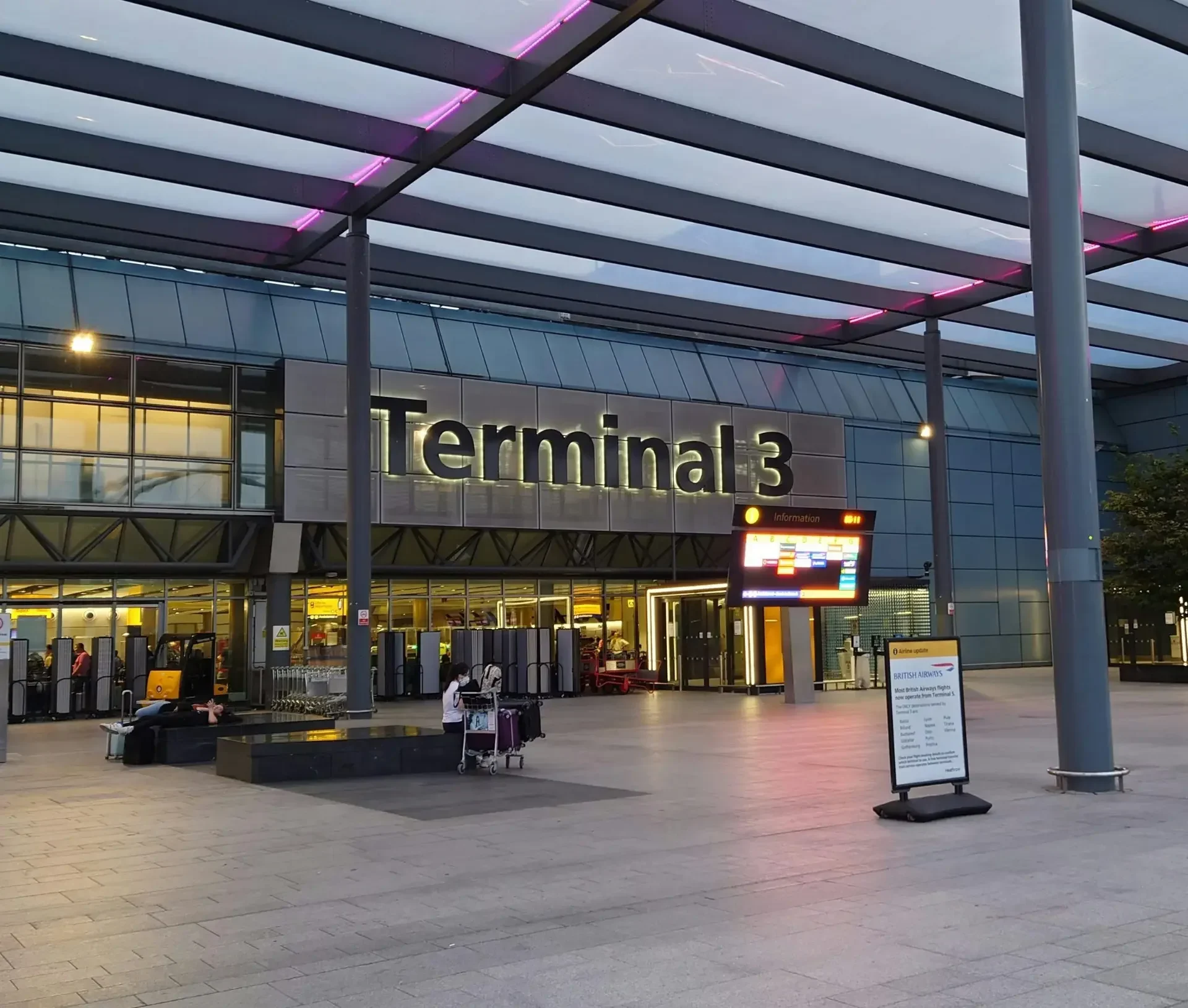 Exterior of terminal 3 at an airport during early evening, with a few people and luggage outside; illuminated signage and digital information boards highlight the heightened cybersecurity measures now common after recent ransomware threats at airports.