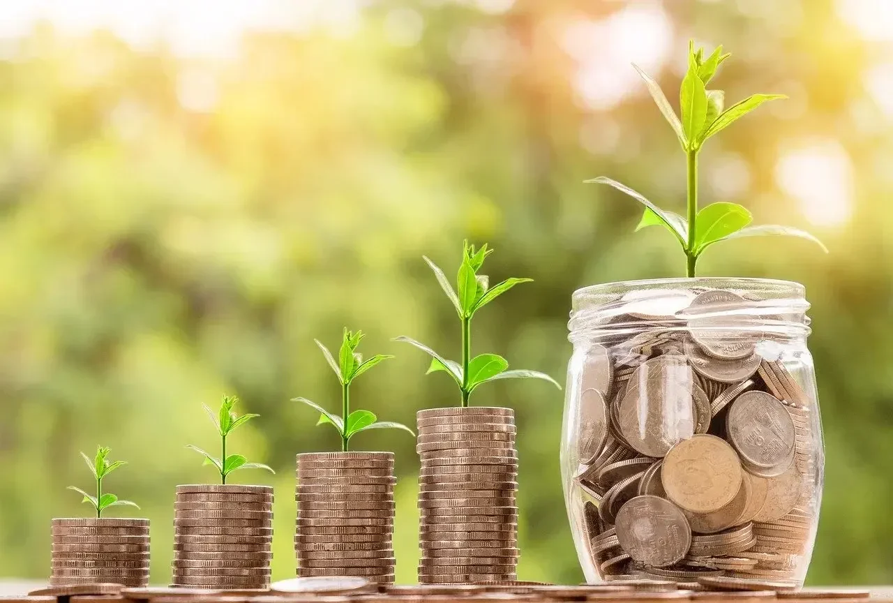 Four stacks of coins and a jar filled with coins, each topped with a growing plant, symbolize financial growth and the it support benefits gained from outsourcing it support. green blurred background.