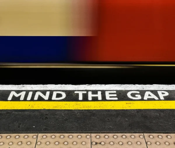 The phrase "mind the gap" is painted in white on a train platform edge with a yellow line, while a blurred train passes by—symbolizing the it skills gap that many companies face when recruiting top talent.