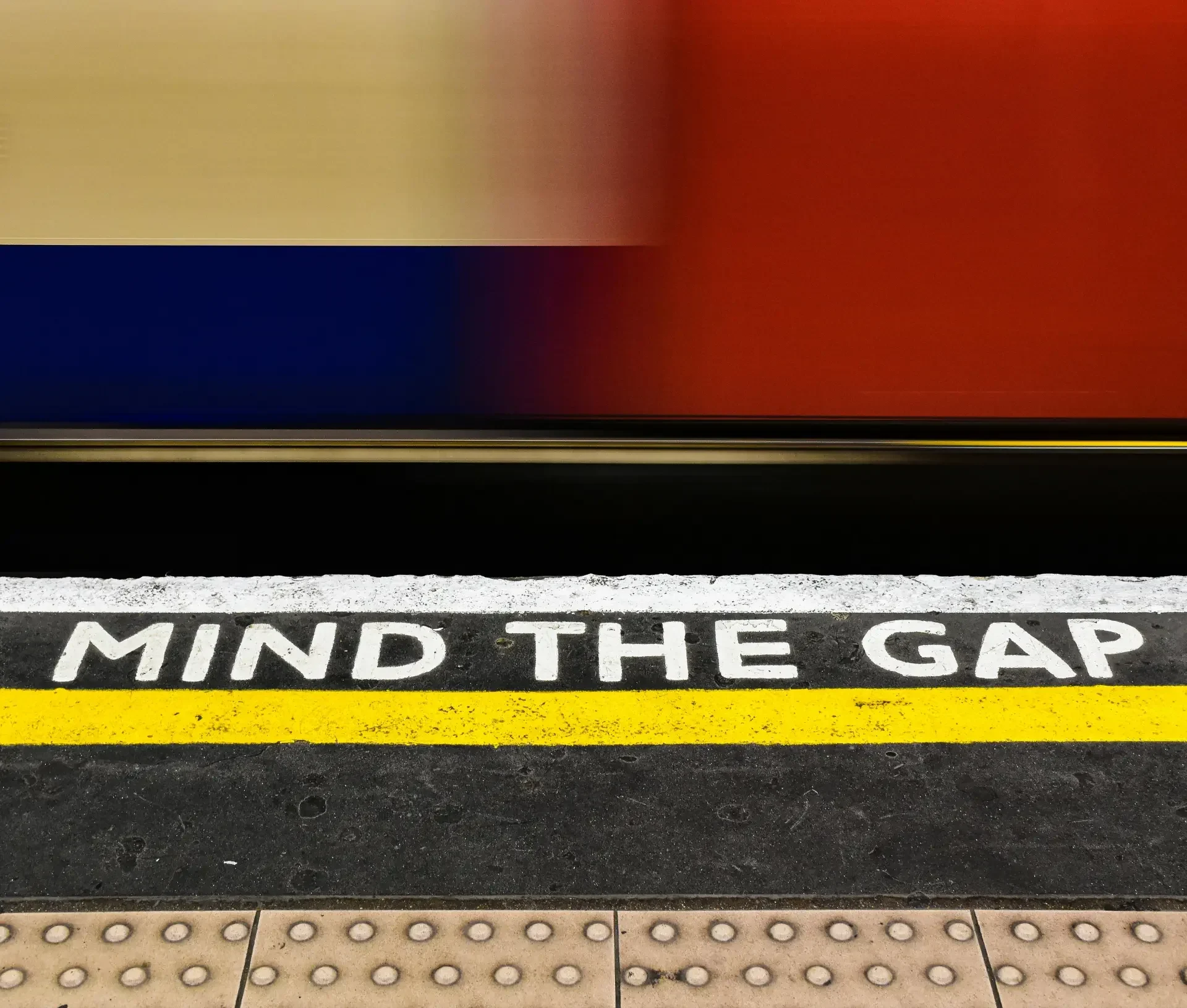The phrase "mind the gap" is painted in white on a train platform edge with a yellow line, while a blurred train passes by—symbolizing the it skills gap that many companies face when recruiting top talent.