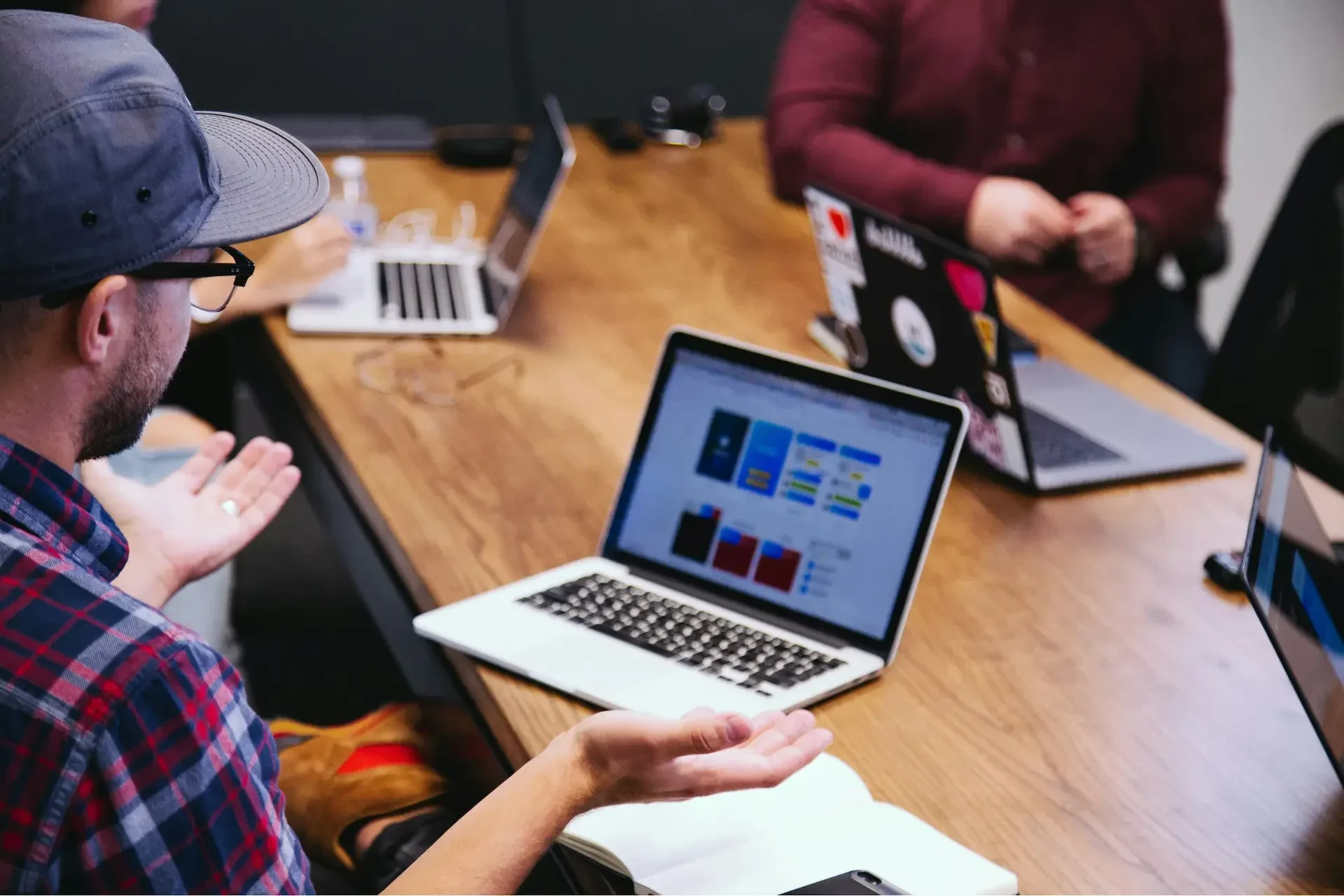 Three people sit at a wooden table with open laptops, engaged in discussion; one person gestures with hands while others focus on their screens.