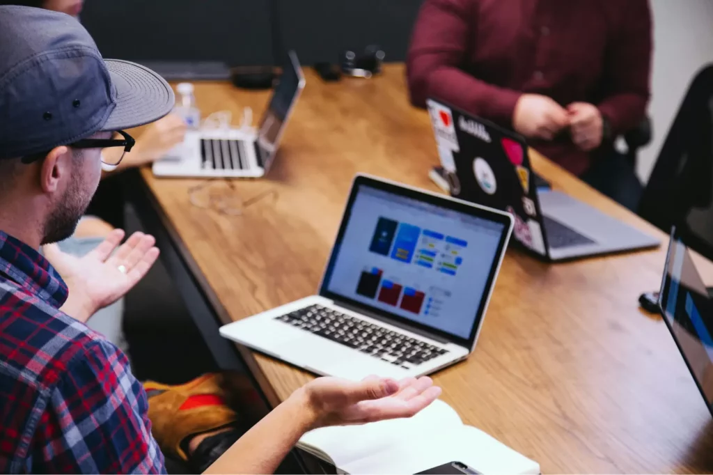 People sitting around a wooden table with laptops open, engaging in a discussion. One person gestures with hands while others appear to listen or work on their computers.
