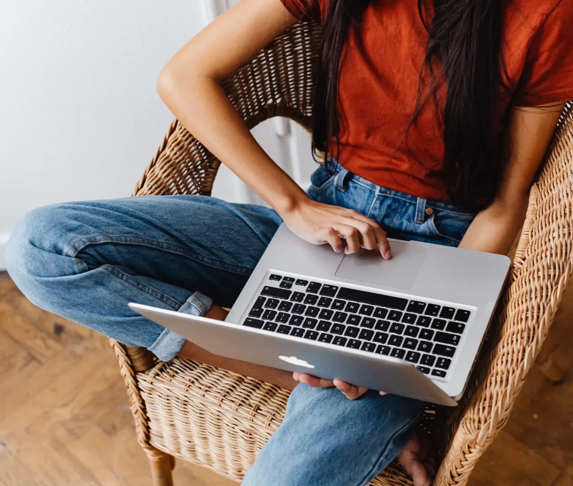 A person sitting in a wicker chair, wearing blue jeans and a rust colored shirt, using a laptop on their lap—working efficiently as part of a remote team.