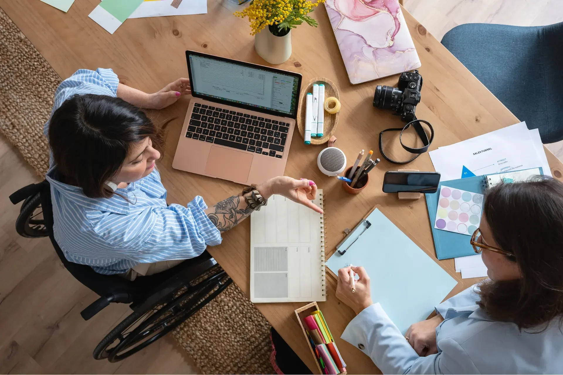 Two women work together at a desk with a laptop, documents, and camera—collaborating on projects for marketing agencies; one woman uses a wheelchair.