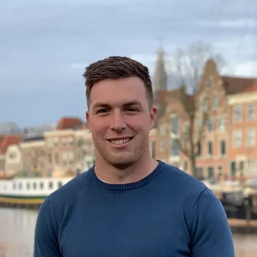 A man wearing a blue sweater stands outdoors in front of a canal with buildings and trees in the background.