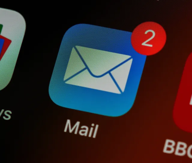 Close up of a Mail app icon with a red notification badge showing 2, indicating two unread messages—highlighting the importance of checking for a phishing email in your workplace. Dark background, other app icons partly visible.