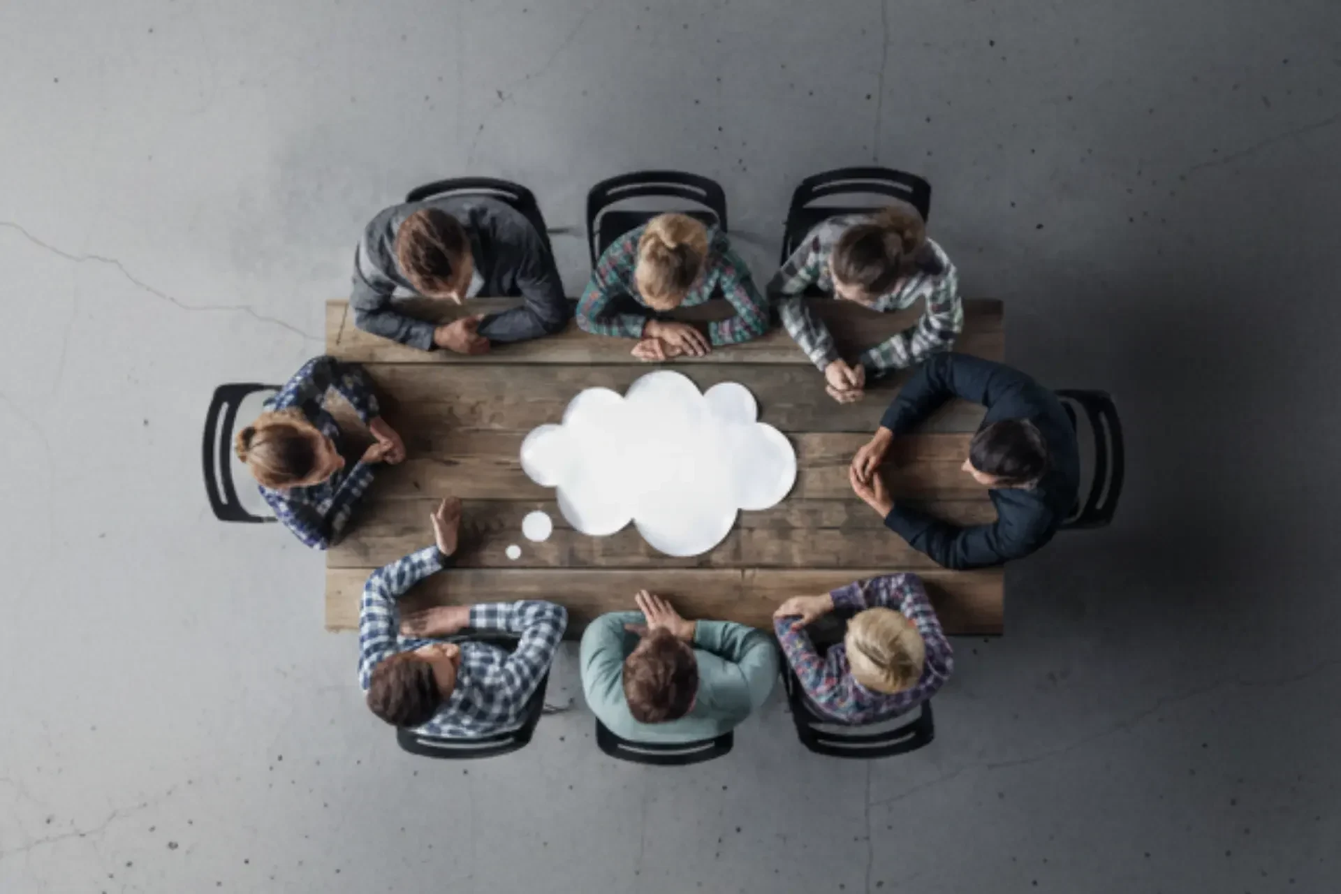Seven people sit around a wooden table with a white thought bubble shape in the center, viewed from above—an ideal setting for a quarterly it review focused on it metrics and strategic outlook.