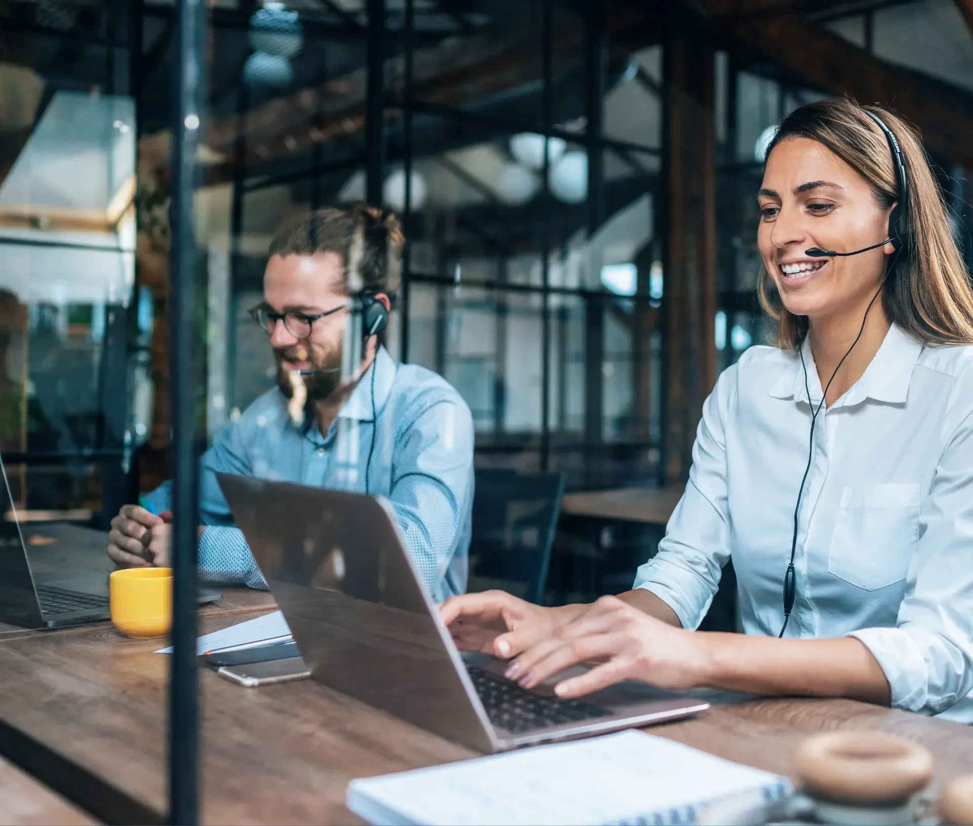 Two people wearing headsets sit at desks with laptops in a modern office, focused on it support or participating in a virtual meeting to boost productivity.