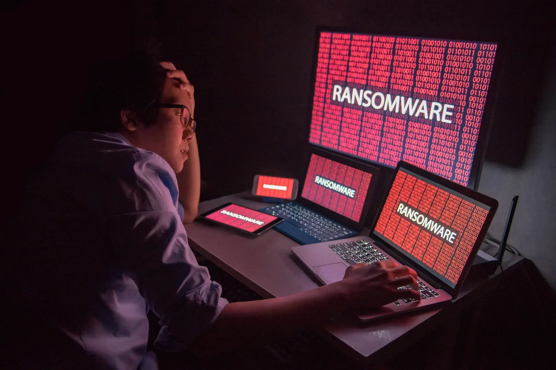 A person sits at a desk, looking stressed, surrounded by multiple screens displaying red ransomware warning messages—highlighting the need for ransomware tips and mac safety to stay protected.