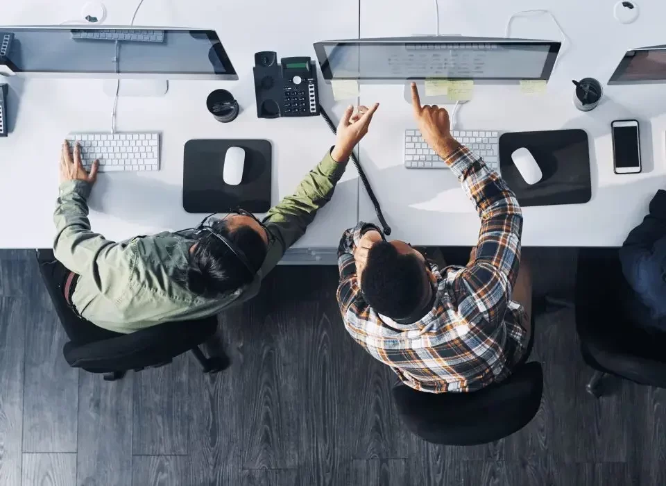Two people seated at a desk working on computers, with the person on the right pointing at the screen. They are wearing headsets and surrounded by office supplies. Top-down view.