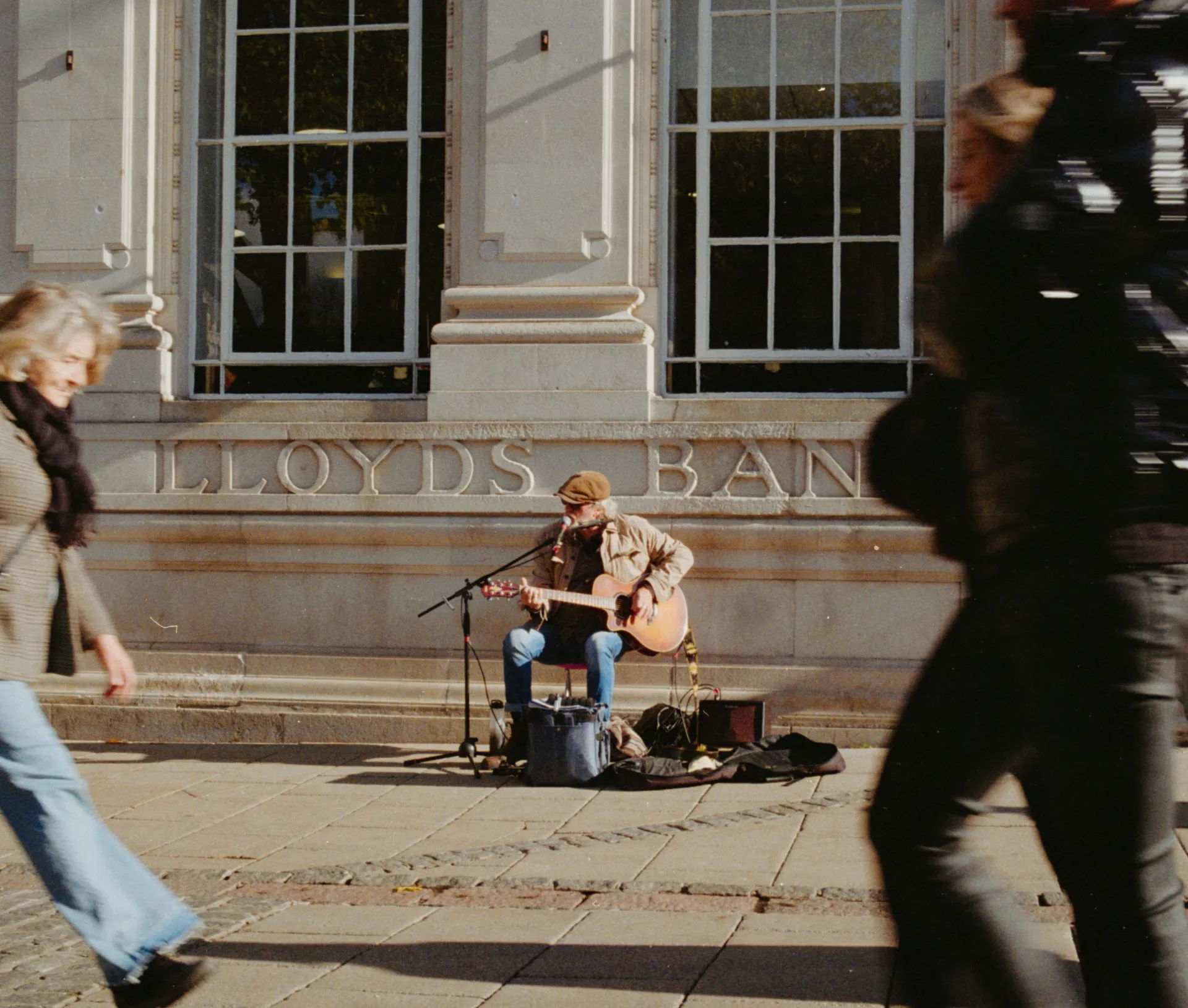 A street musician sits and plays guitar outside a building labelled “Lloyds Bank,” a recognisable name in regulated industries, as people walk by on the pavement, appearing blurred due to motion.