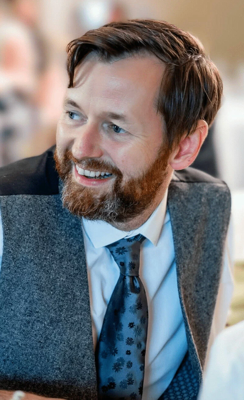 Bearded man in a gray vest and blue patterned tie smiling, looking to the side.