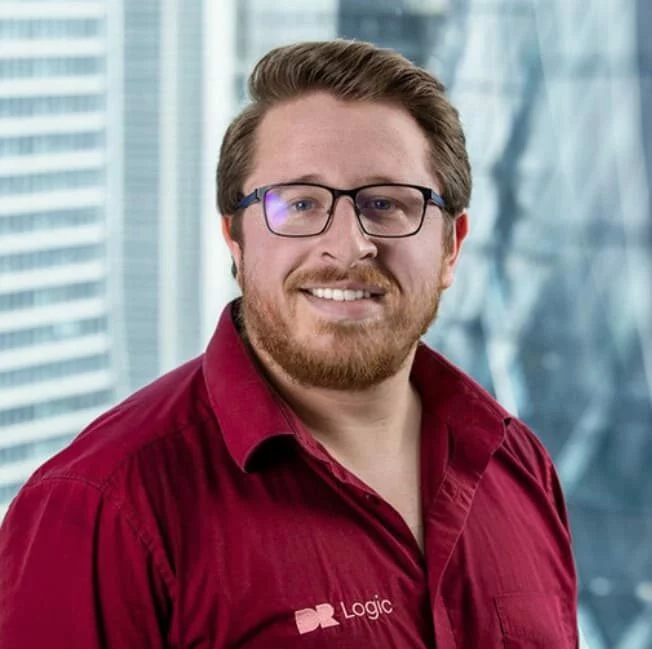 Man with glasses and a beard wearing a red shirt stands in front of a window overlooking a cityscape.