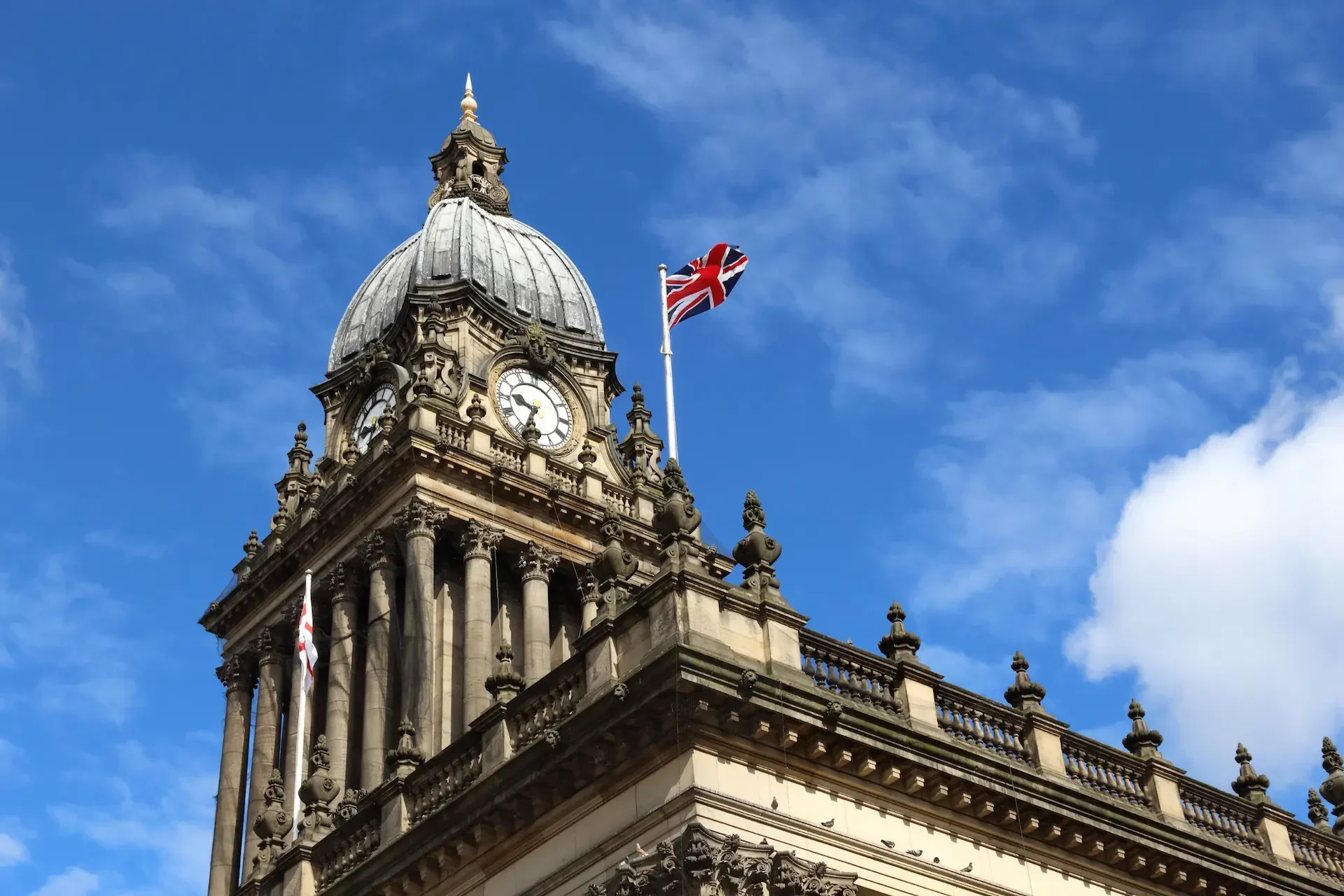 Clock tower of a historic stone building with a domed roof, columns, and a union jack flag flying atop, standing as a symbol of secure innovation and government funding, set against a blue sky with scattered clouds.