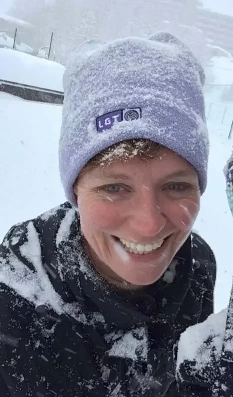 A person in a light purple beanie and black jacket smiles outdoors, with snow covering their face, hat, and clothing.