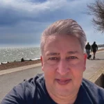 A person takes a selfie on a sunny day by the seaside. the ocean and sky are visible in the background, and a few people are walking along the path.